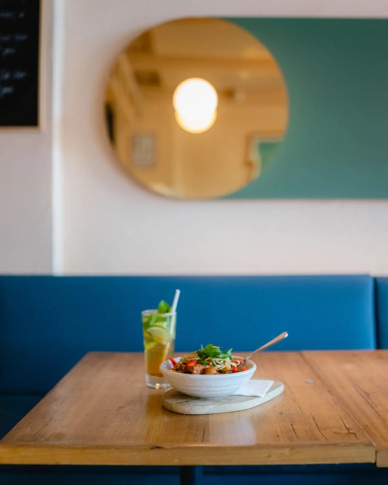Inside Mr Frankie: a blue banquette, a round mirror and a bowl of food with a drink on a timber table